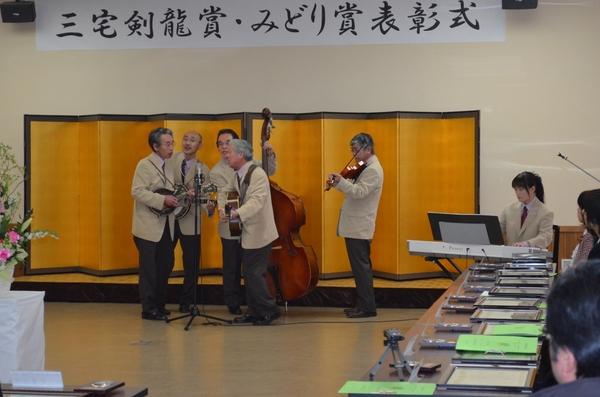 金屏風の前右側でエレクトーンを弾いてる女性とギターやバイオリン、コントラバスを手にして一本のマイクを囲んで演奏しているフォークソンググループのメンバー男性5名の写真